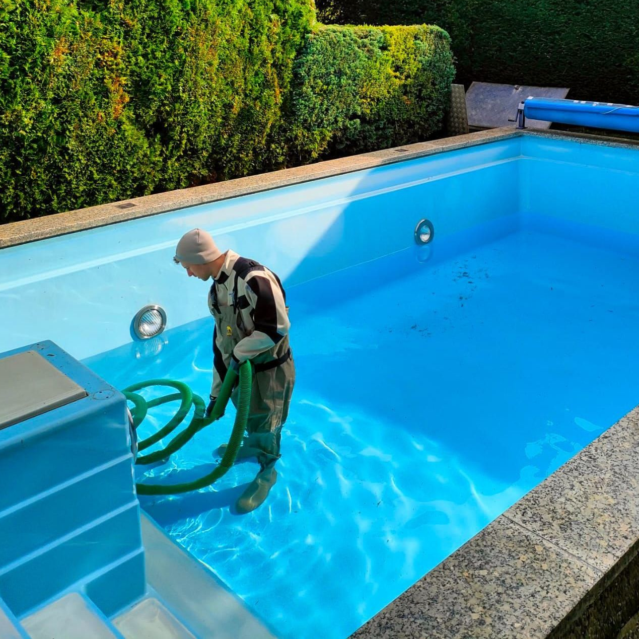 Eine Person reinigt mit einem Gartenschlauch einen leeren blauen Swimmingpool im Hinterhof.