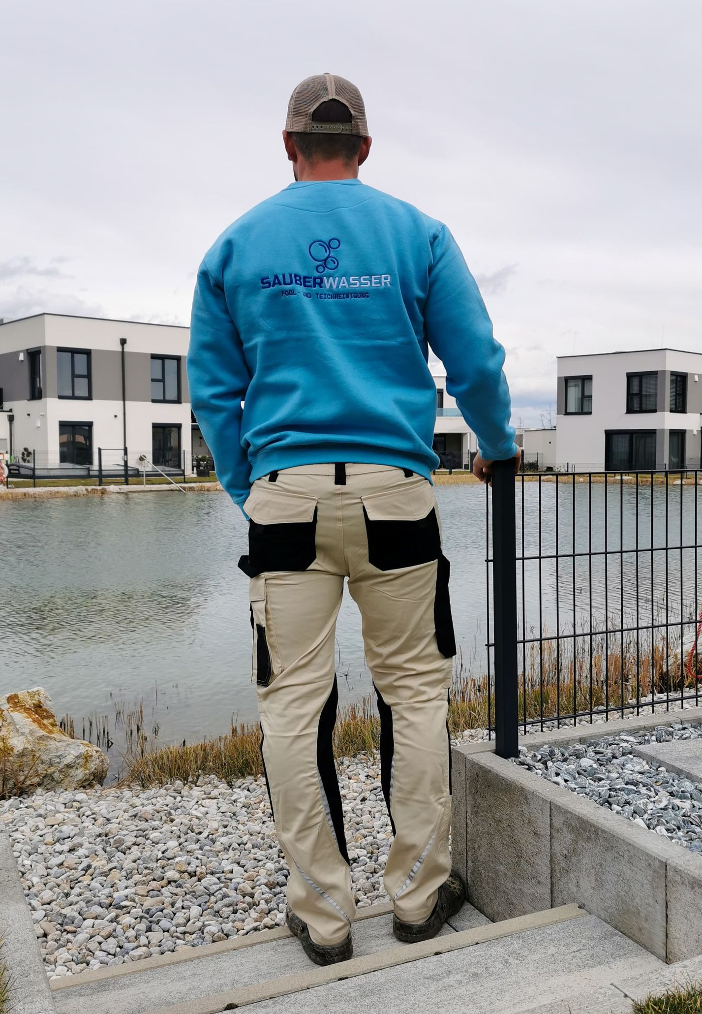Eine Person in blauem Sweatshirt und beiger Hose steht an einem See, blickt in Richtung moderner Häuser.