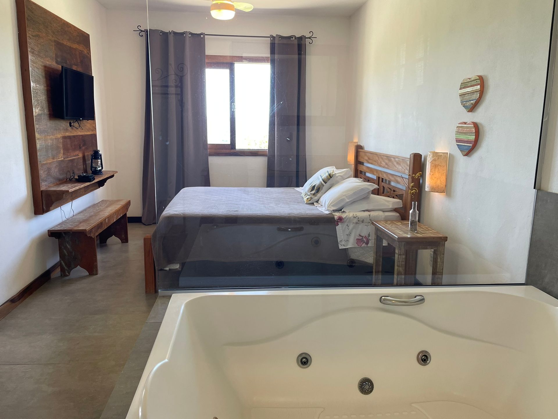 Um quarto com uma grande banheira de hidromassagem em primeiro plano, apresentando uma cama, parede com detalhes em pedra e um banco de madeira.
