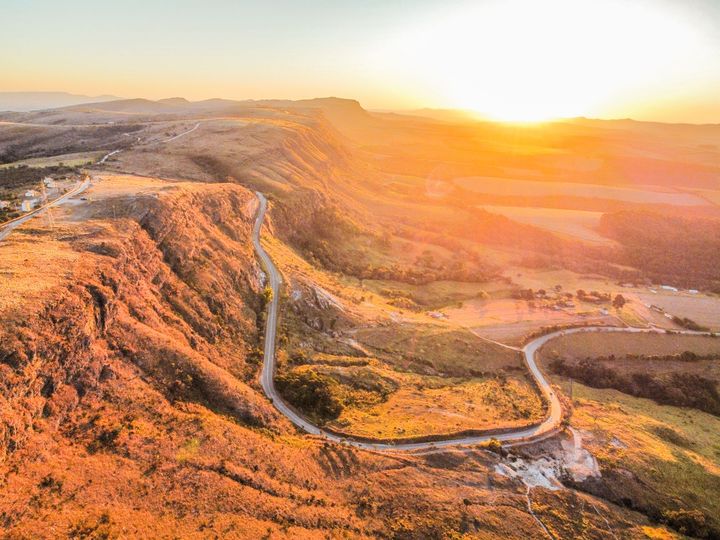 Ao pôr do sol, uma estrada sinuosa serpenteia pela beira de um penhasco acidentado, lançando um brilho dourado e quente sobre a paisagem montanhosa.