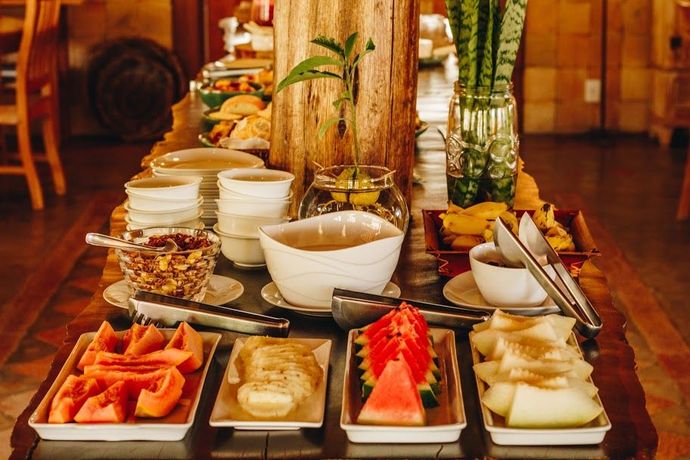 Uma mesa de buffet de café da manhã com pratos de frutas fatiadas, tigelas pequenas, pilhas de louça e um vaso de folhas verdes.