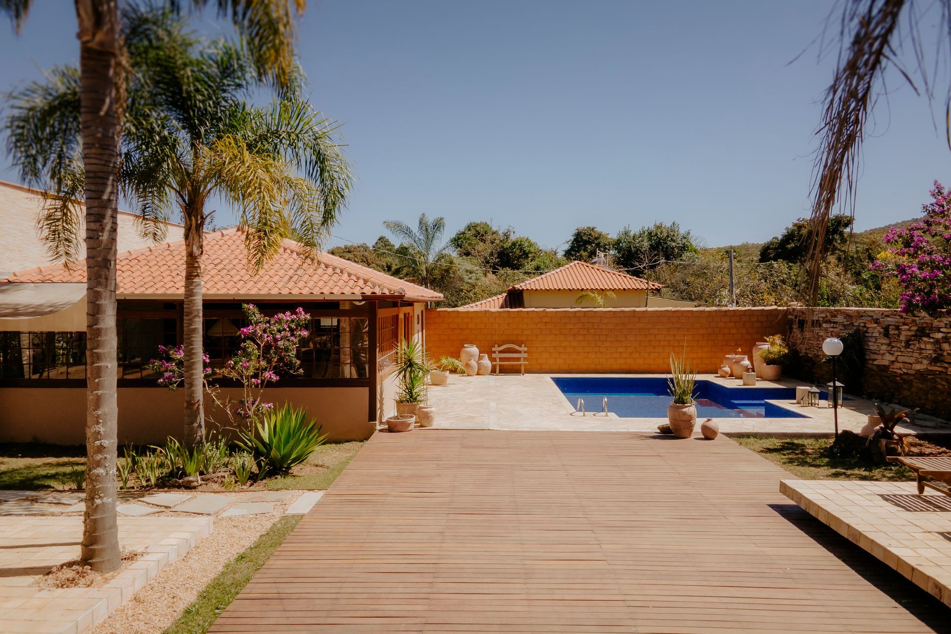 Um deck de madeira leva a uma piscina retangular ao lado de uma casa com telhado de telhas de terracota, sob um céu azul claro.