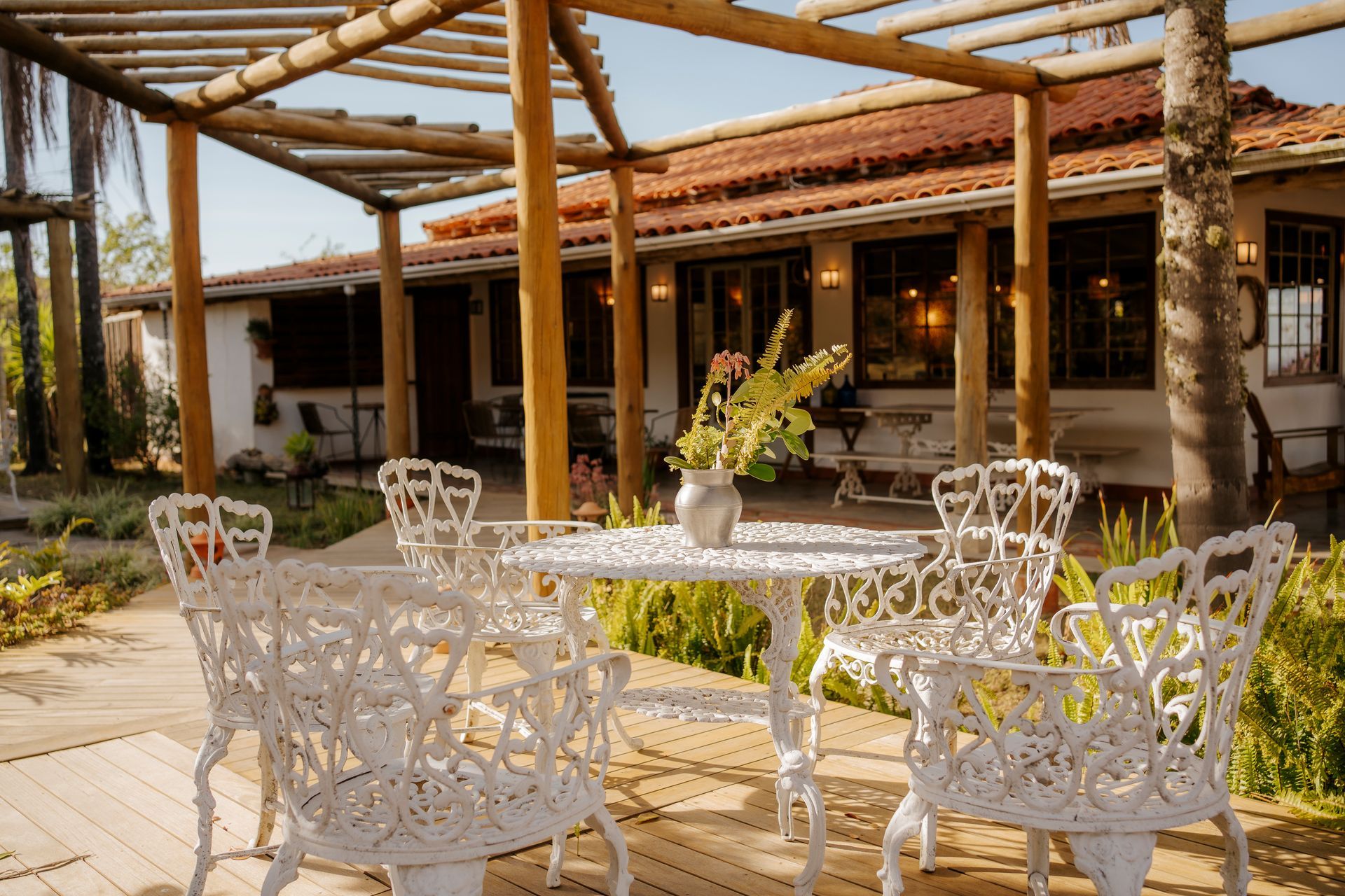 Uma mesa e cadeiras de ferro branco estão dispostas em um pátio sob uma pérgola de madeira em frente a uma casa rústica de cor clara.