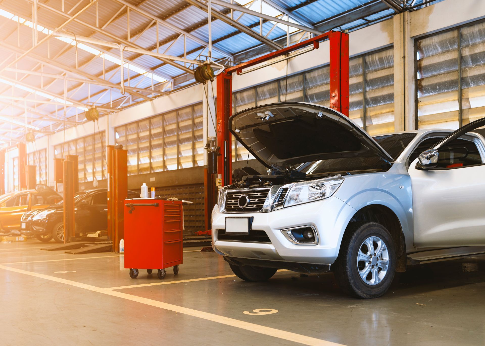 Two silver SUVs in an auto repair shop, one with hood open beside a red tool cart.