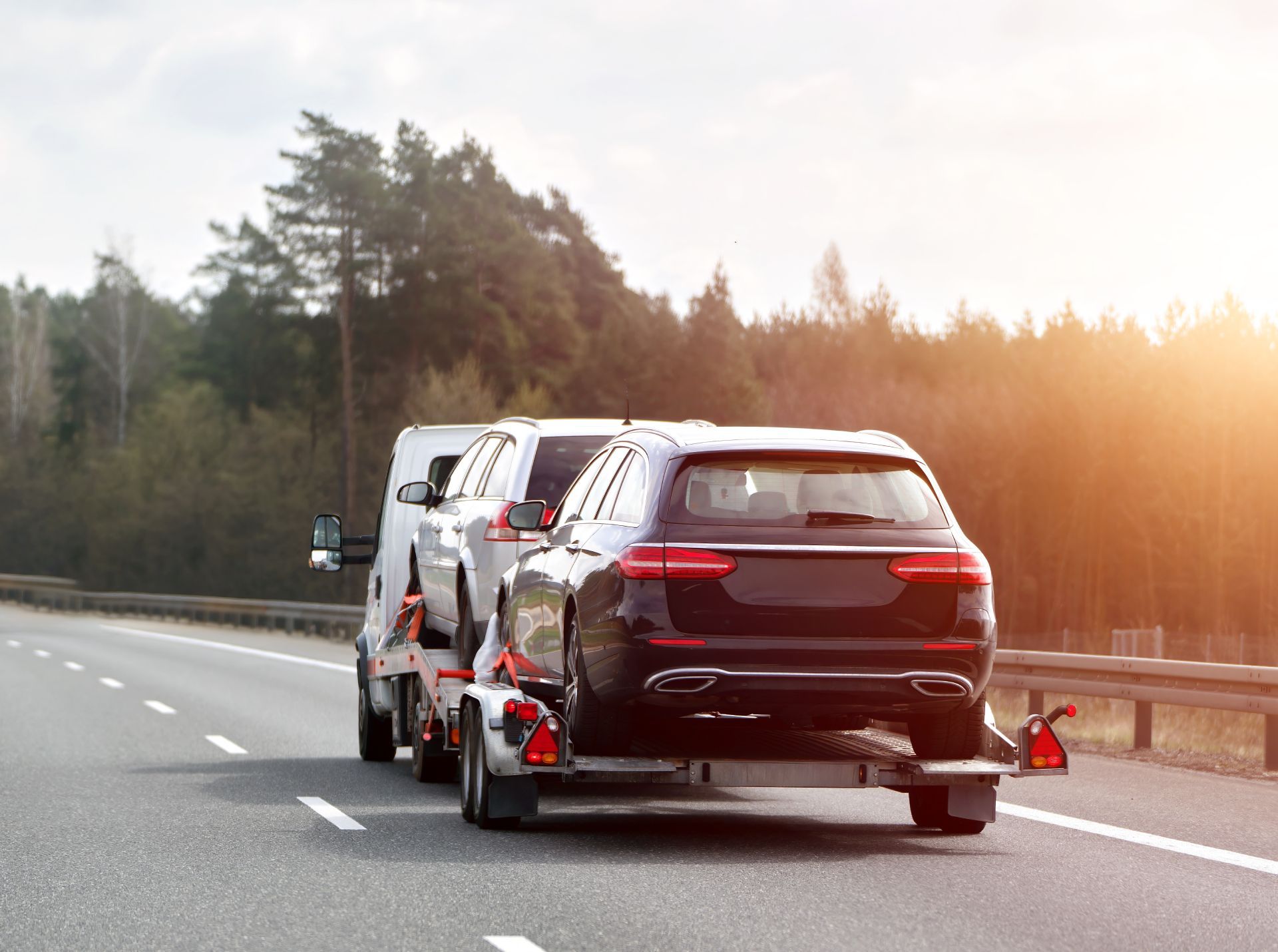 Car towing a white van on a highway at sunset, with orange cones marking the tow trailer.