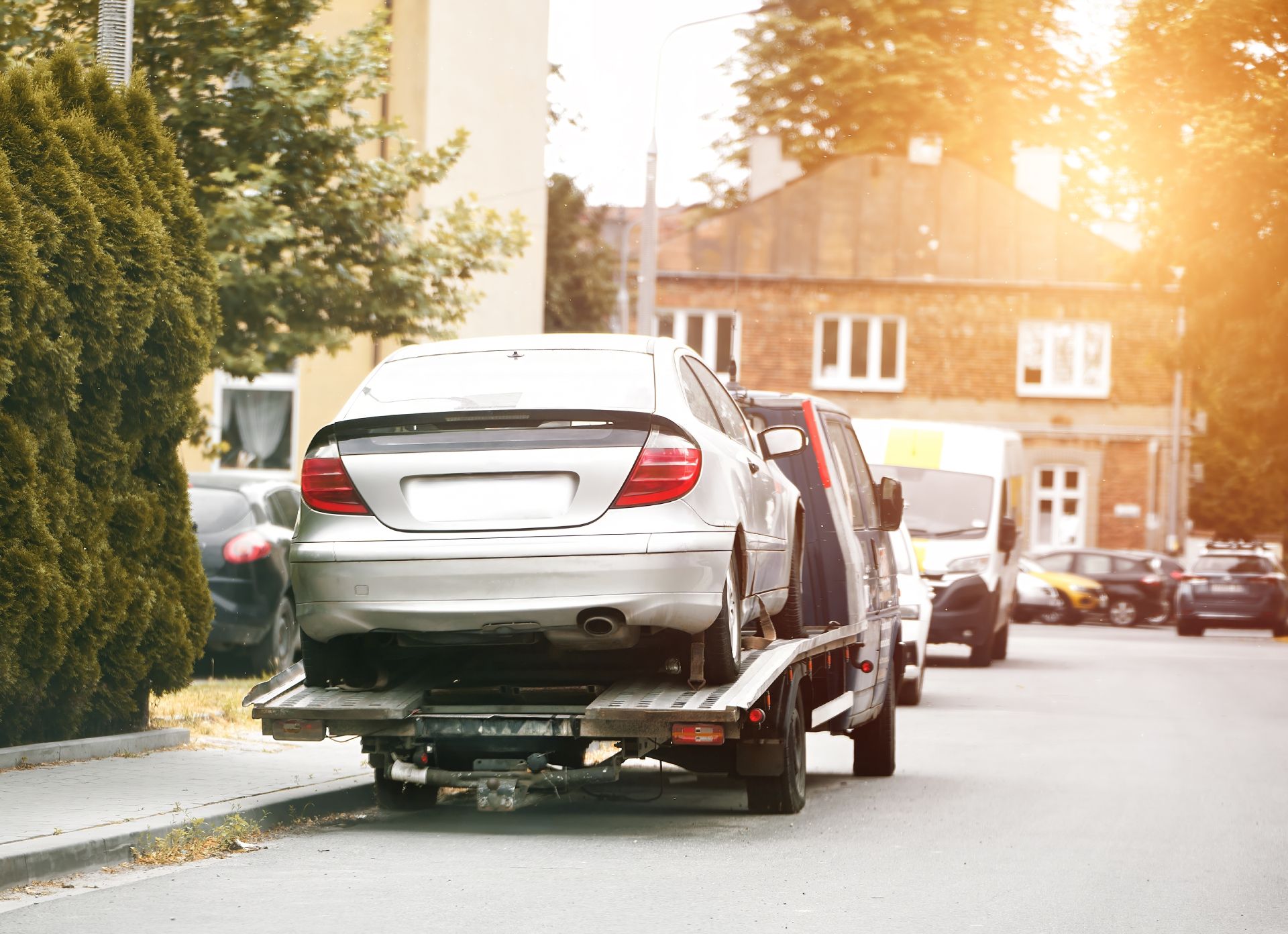 Silver sedan loaded on a tow truck on a sunlit street, with houses in the background