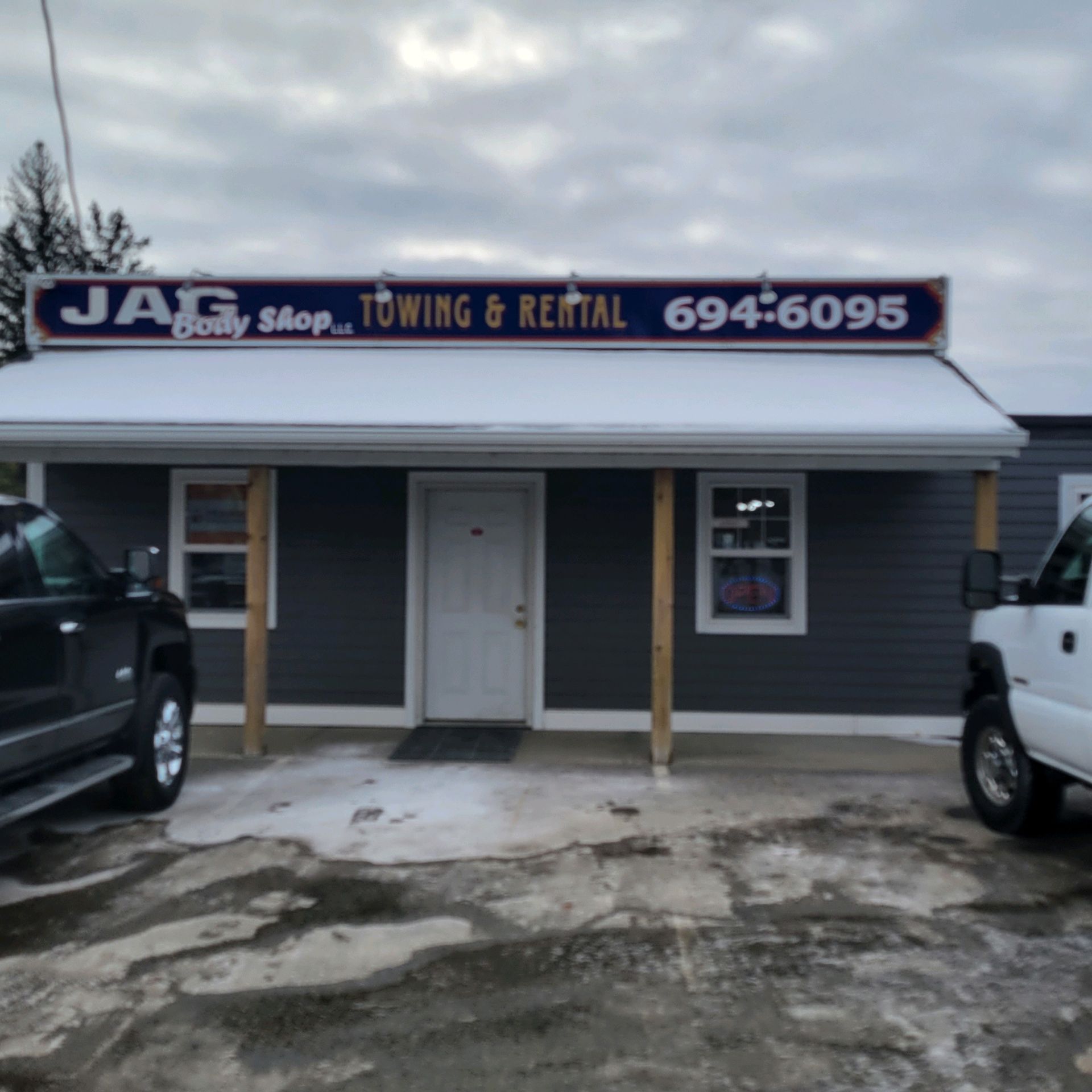 JAG Body Shop towing and rental storefront with gray exterior and parked trucks under a cloudy sky