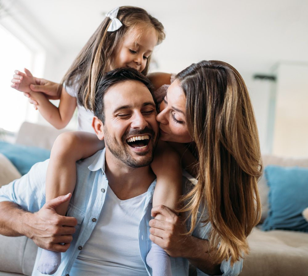 Family laughing together: daughter on dad's shoulders, mom kissing dad's cheek, in a living room.