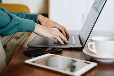 Hands typing on a laptop beside a smartphone, tablet, and white coffee cup on a table