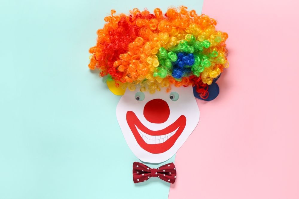 Colorful clown face with rainbow curly wig, red nose, smiling mouth, and red bow tie on pastel background