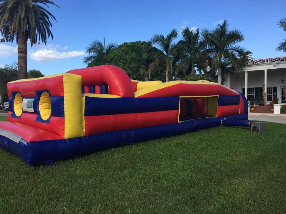 Colorful red, yellow, and blue inflatable obstacle course on a grassy lawn with palm trees and buildings behind it