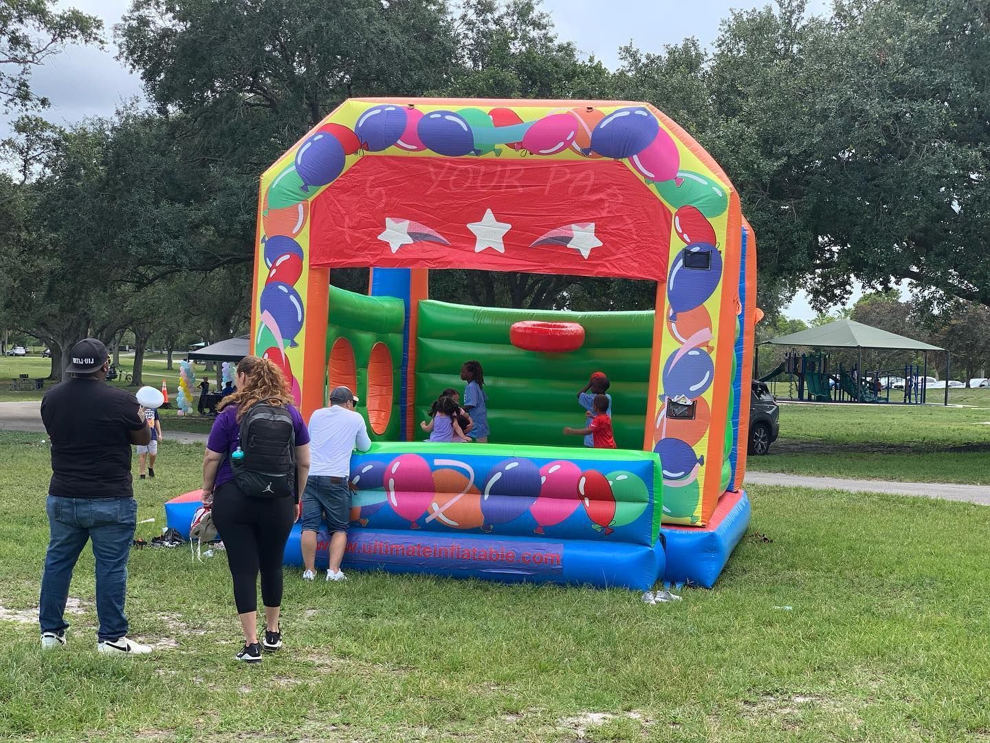 Colorful inflatable bounce house at a park with children inside and two adults standing nearby