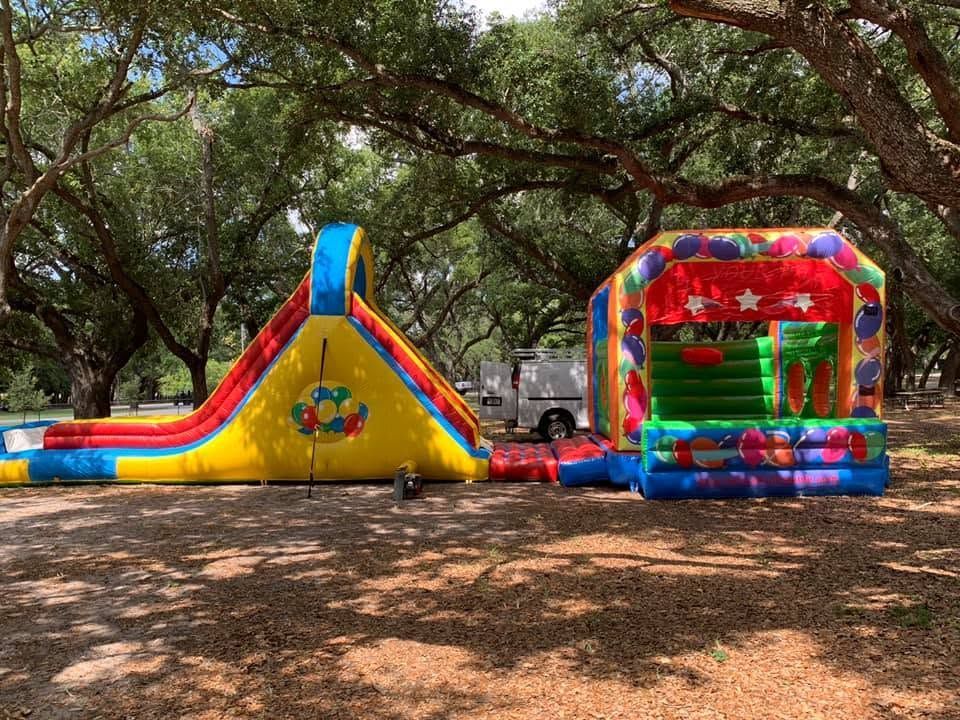 Colorful inflatable slide and bounce house at a park under large trees