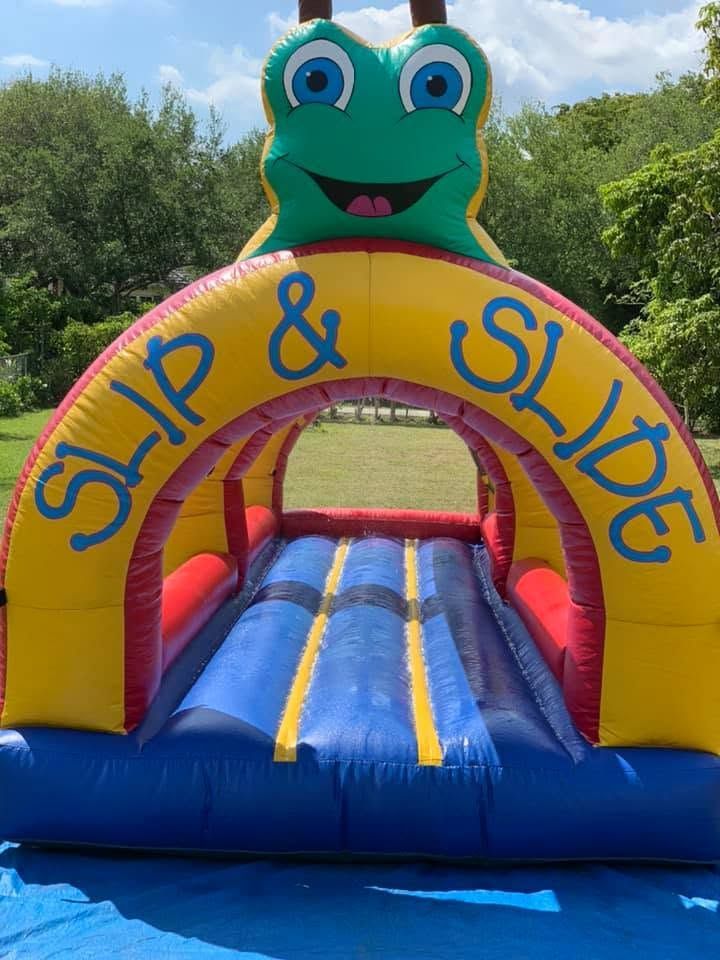 Colorful frog-themed “Slip & Slide” inflatable bounce slide on grass under a blue sky