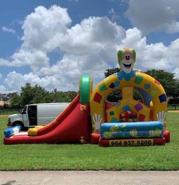 Inflatable clown bounce house and red slide on a grassy lawn under a cloudy sky