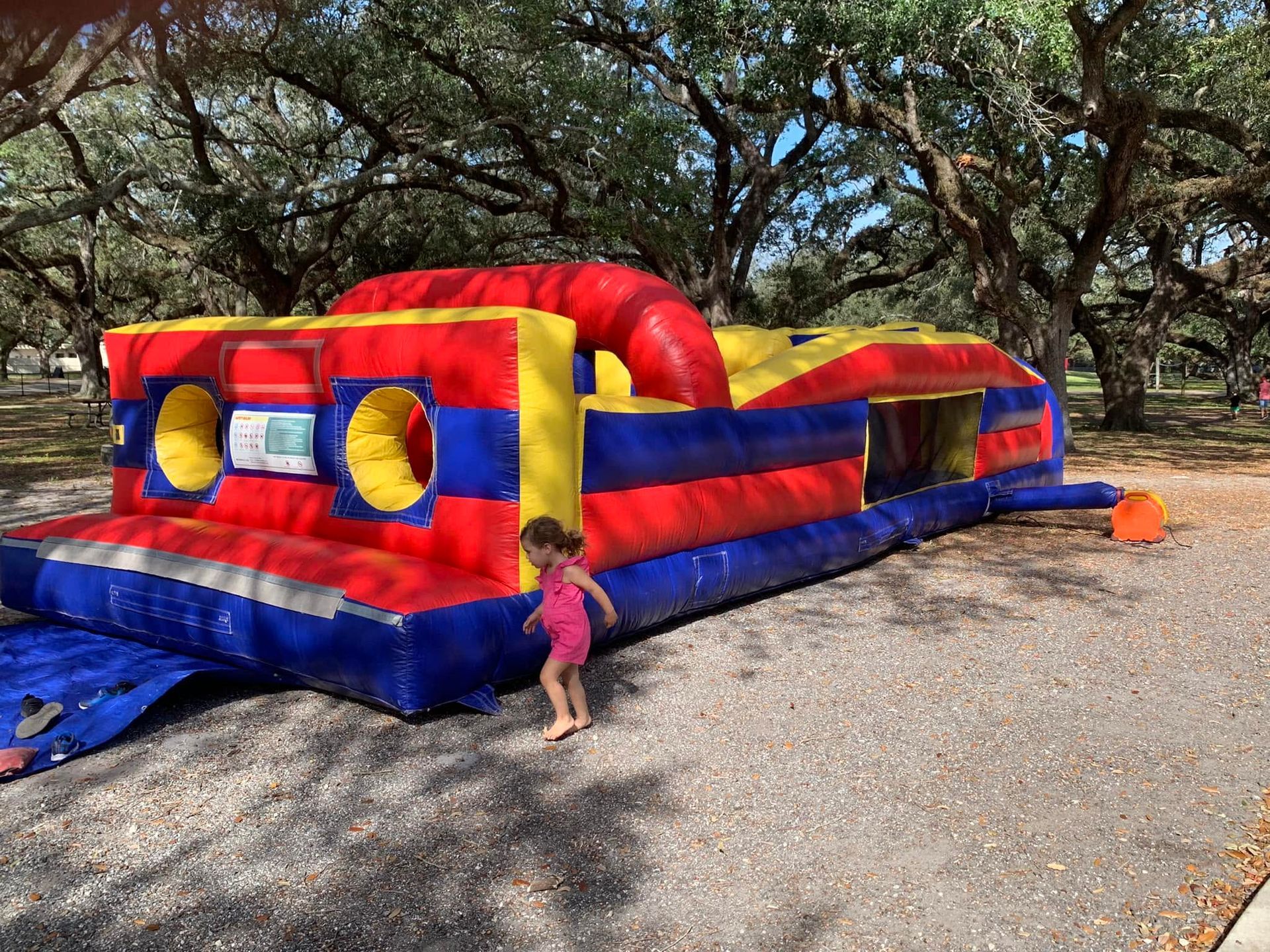 Colorful red, yellow, and blue inflatable bounce house on gravel with a child standing beside it under trees