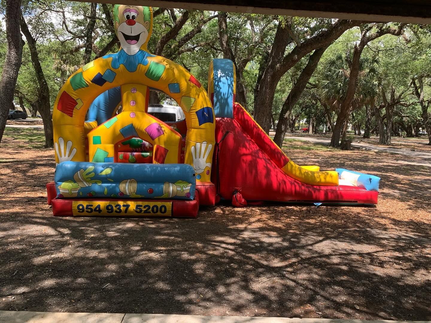 Colorful clown-themed bounce house and slide in a shaded park, with a large clown face at the entrance.