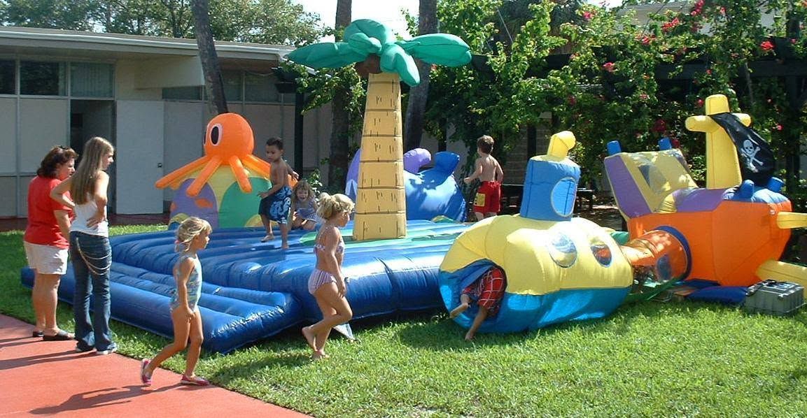 Children playing on a colorful inflatable water slide with palm tree decorations in a sunny yard