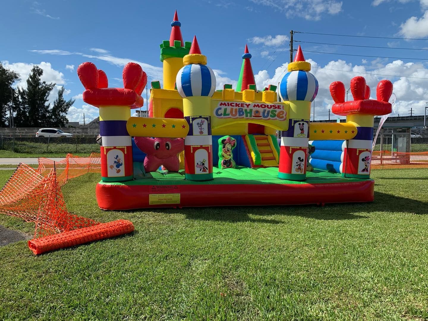 Colorful inflatable bounce house on a grassy field with orange safety netting and a blue sky.