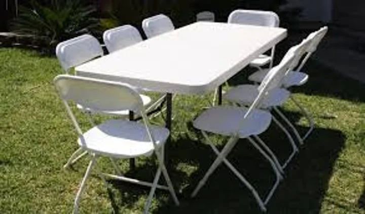 White folding table surrounded by eight white folding chairs on a grassy lawn outdoors