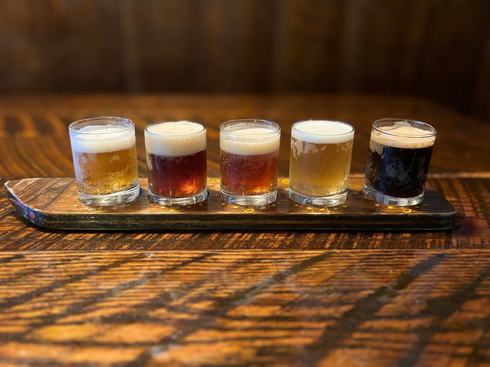 Five glasses of beer are lined up on a wooden tray on a wooden table.