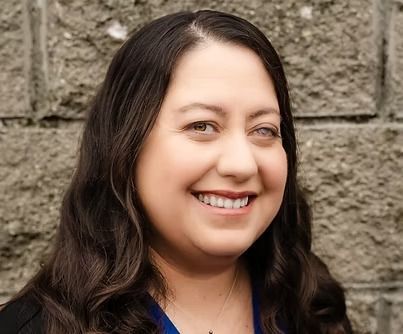 A person with long, dark hair smiles warmly in front of a textured gray stone wall.