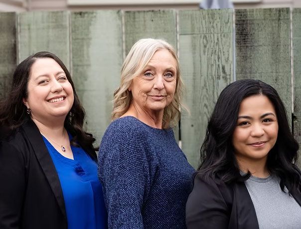 Three women standing in front of a weathered wooden fence; one woman smiles while the others are more neutral.