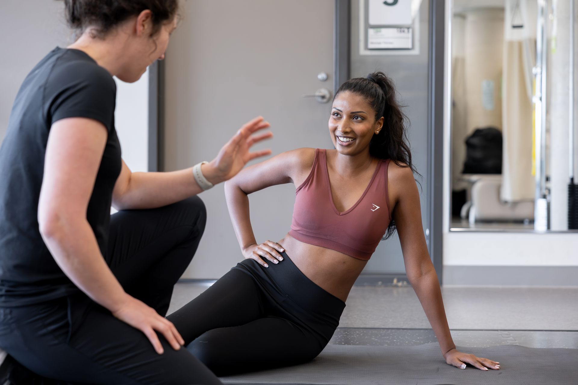 A woman is helping another woman do exercises on a mat in a gym.