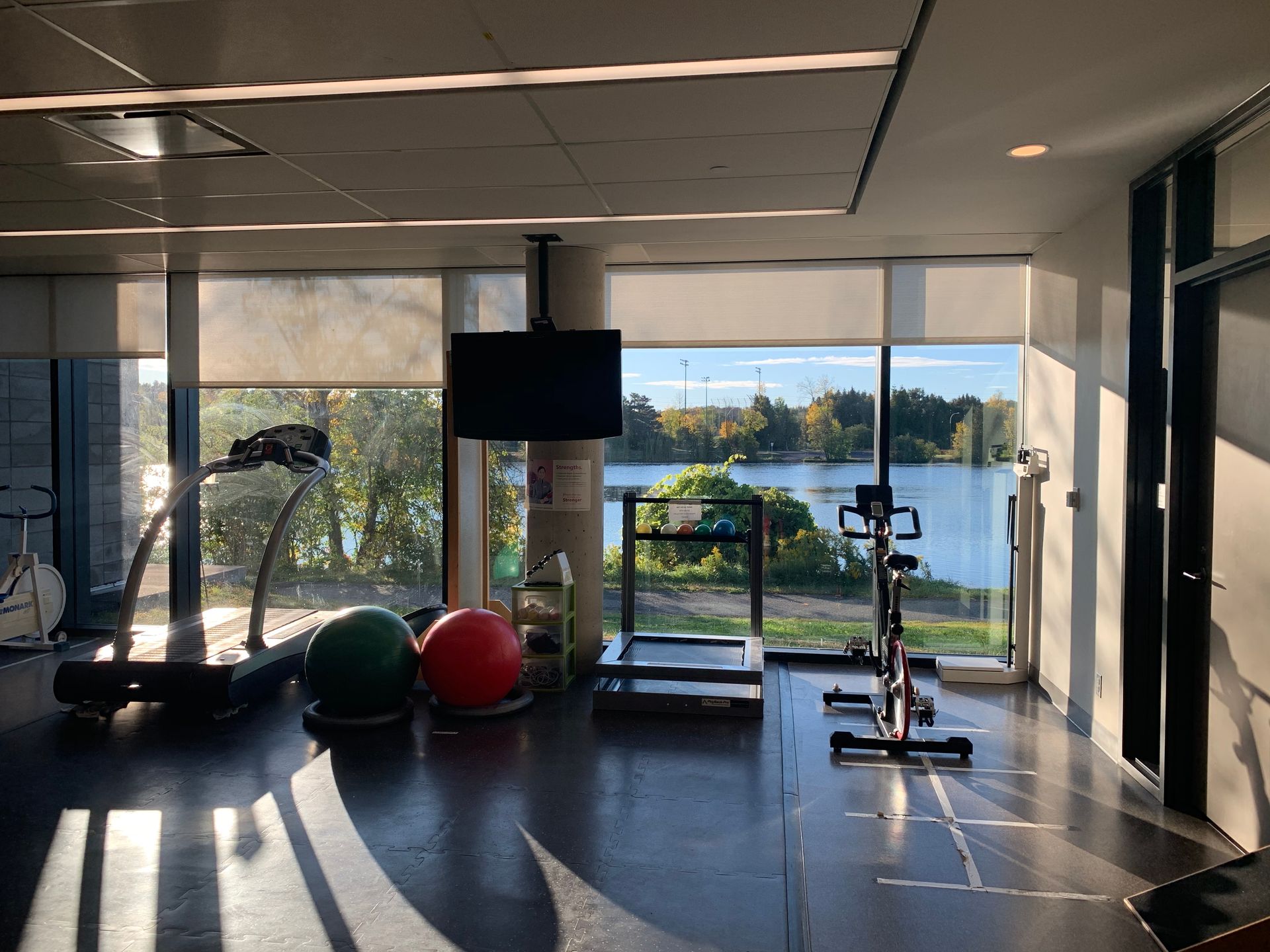 Gym with exercise equipment, windows overlooking water, a sunny day.