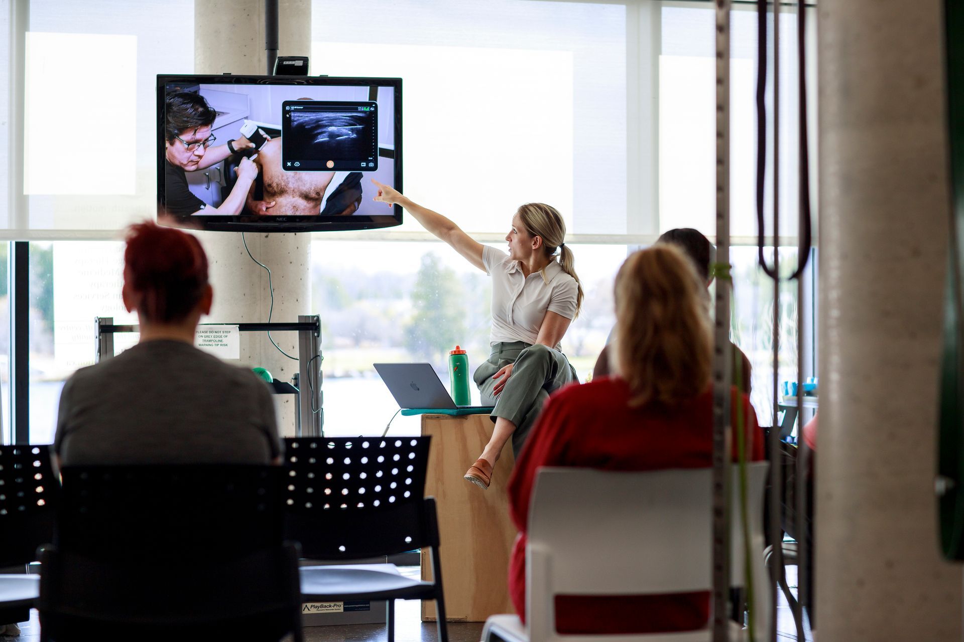 Presenter pointing at a screen during a meeting with seated attendees in a bright conference room