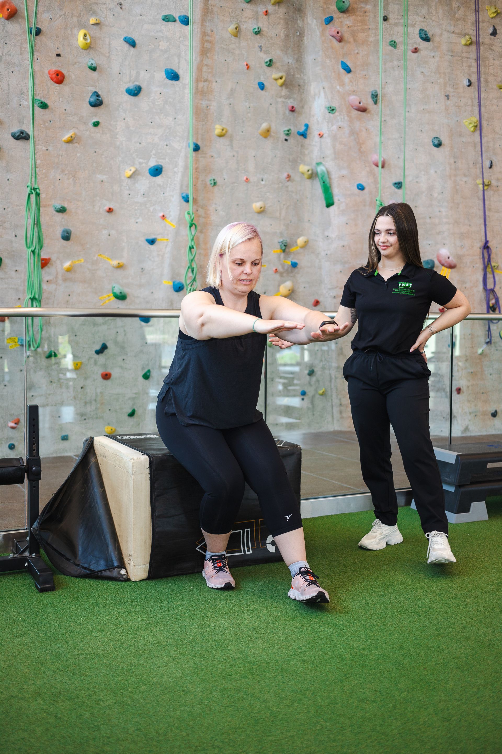 A woman is squatting on a bench in a gym while a woman stands behind her.