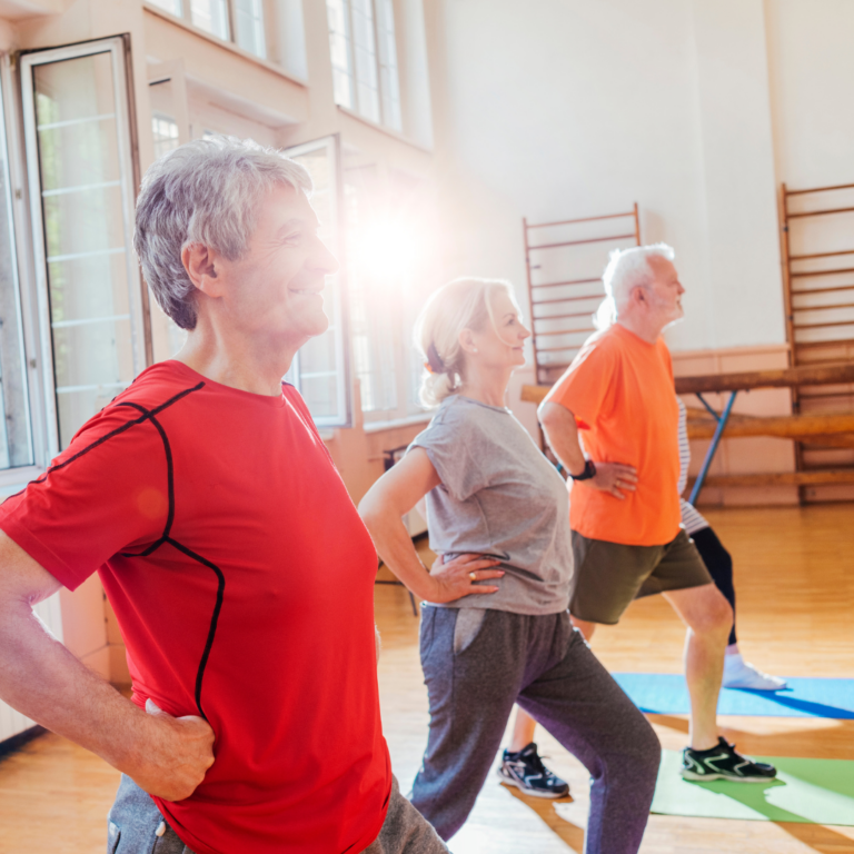 Three older adults in workout attire doing lunges in a gym. Sunlight streams in.
