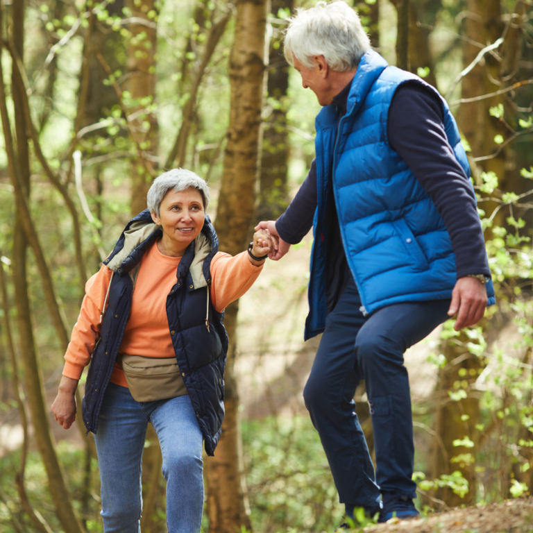 Older couple hiking in a forest; man helping woman ascend a hill.