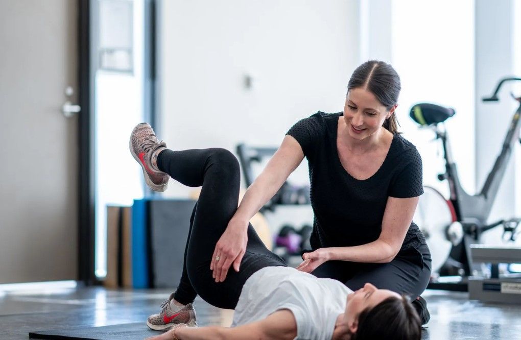 A woman is helping another woman do exercises on a mat in a gym.