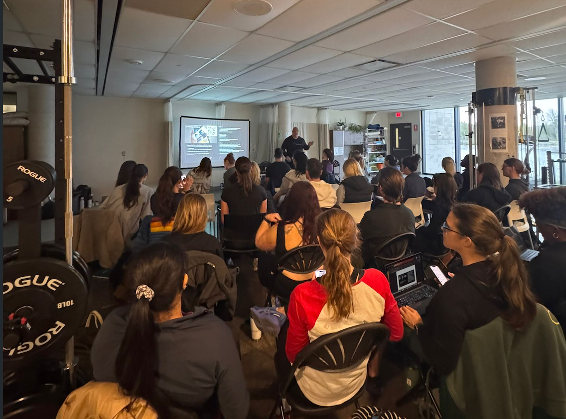 Crowded indoor event with seated audience facing a presentation screen and speaker at the front