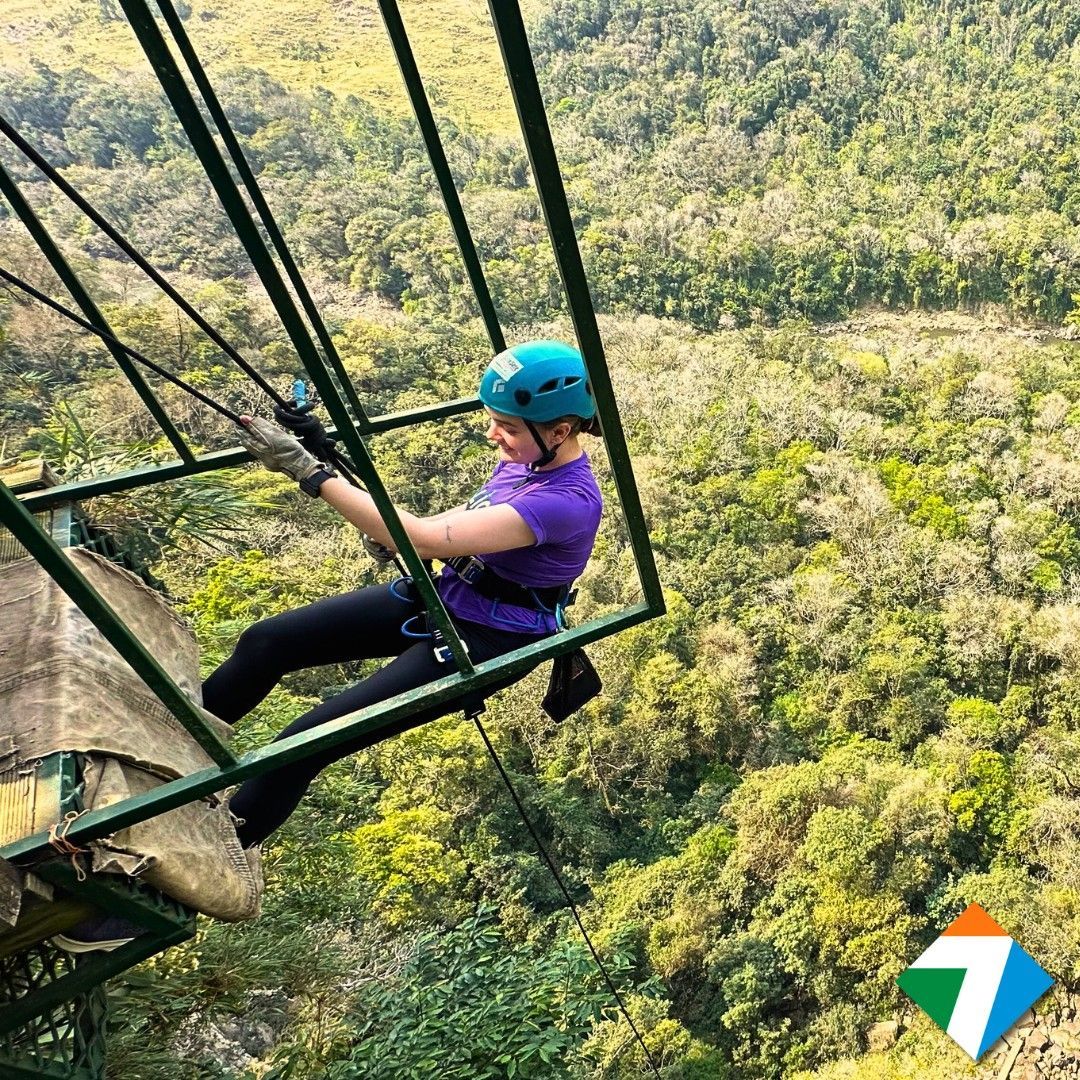 Pessoa em um balanço sobre uma floresta exuberante. Usando capacete, ela se segura na estrutura de suporte.