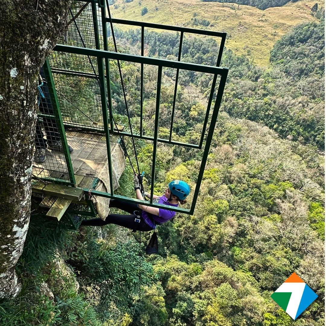 Pessoa descendo de rapel um penhasco dentro de uma gaiola, com uma exuberante floresta verde abaixo.