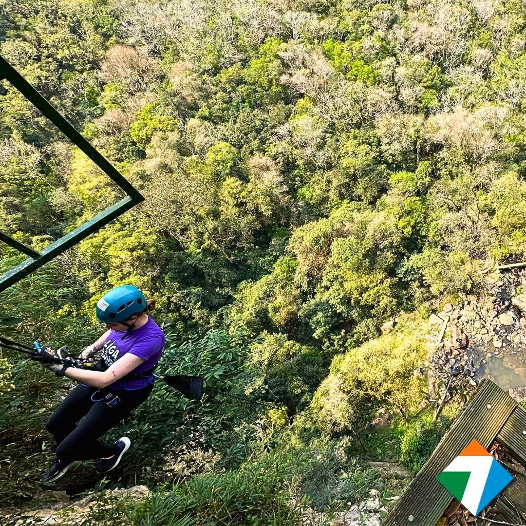 Pessoa deslizando em tirolesa sobre árvores verdejantes, usando capacete e camisa roxa.