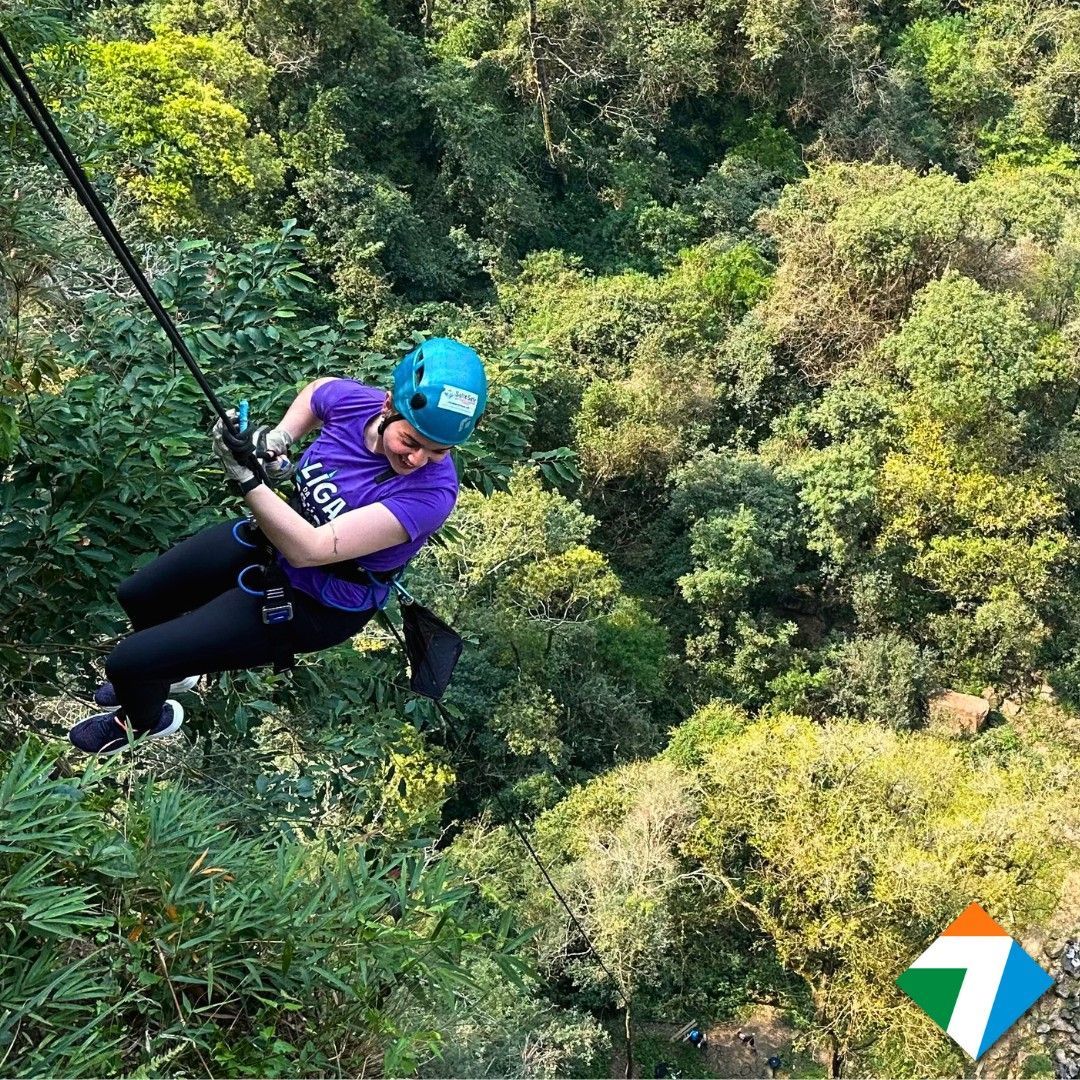 Pessoa deslizando em uma tirolesa por uma floresta verdejante, usando um capacete azul e uma camisa roxa.