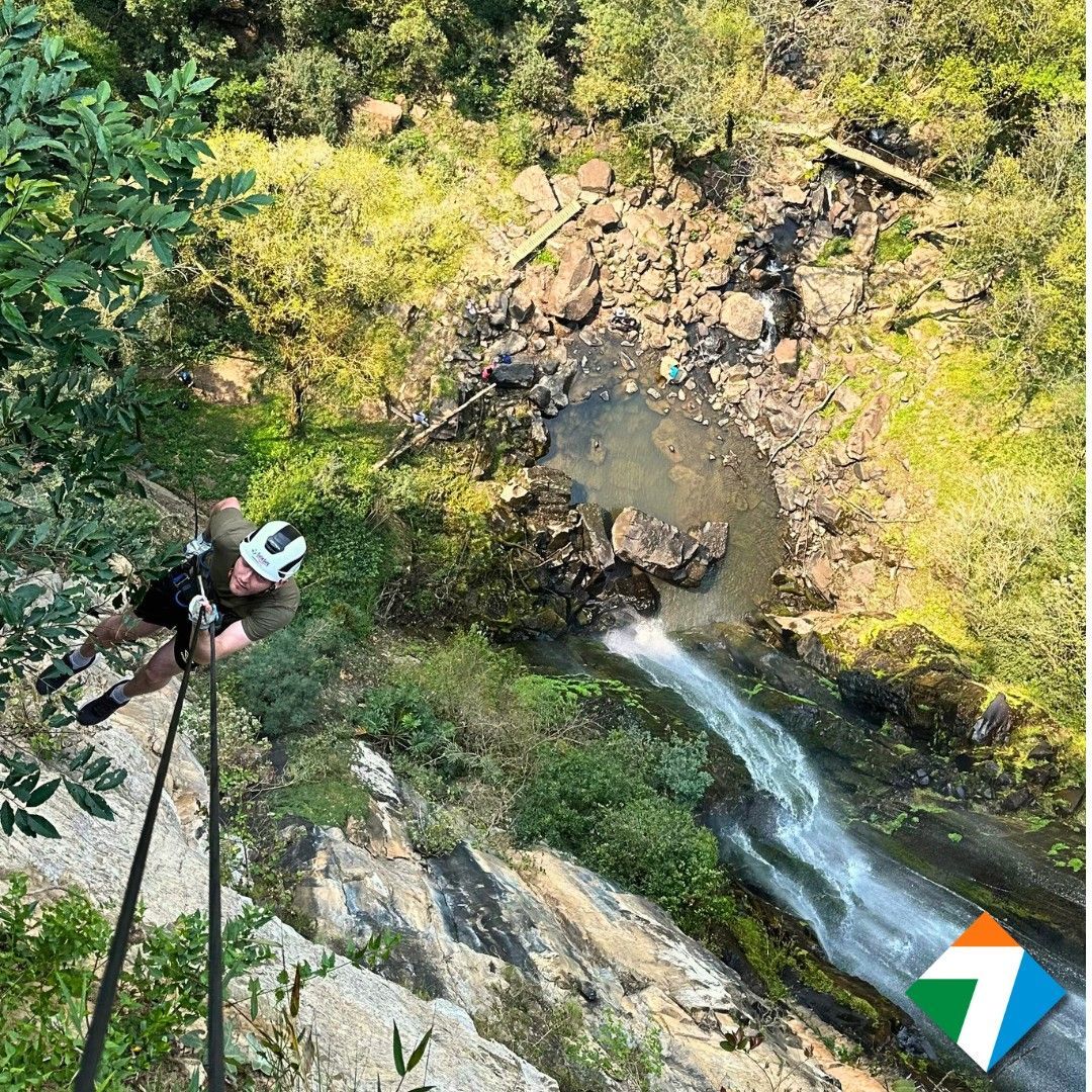 Pessoa fazendo rapel em um penhasco perto de uma cachoeira e folhagem verde.