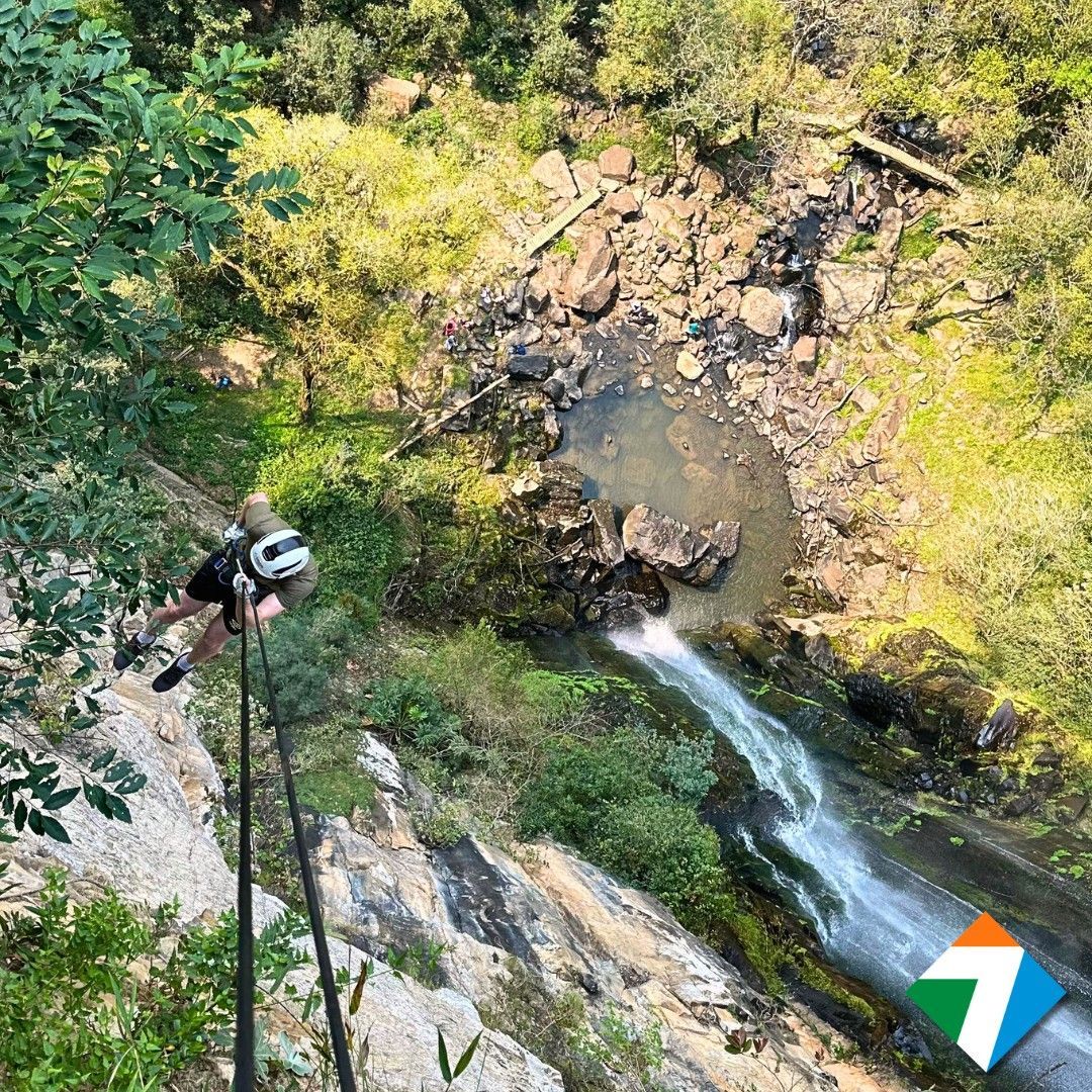 Uma pessoa desce de rapel um penhasco ao lado de uma cachoeira, cercada por árvores verdes e rochas.