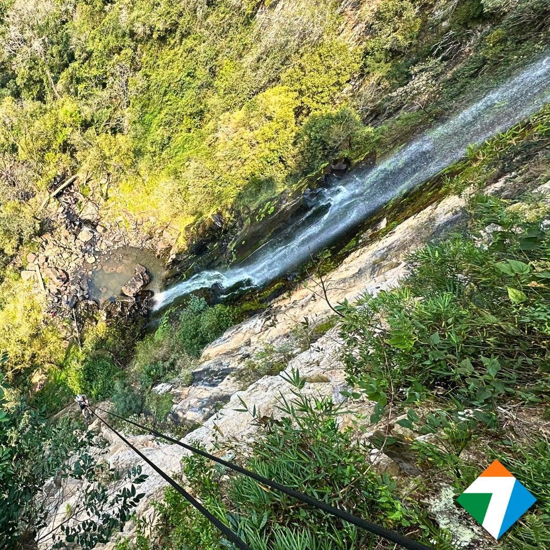 Cachoeira despencando de um penhasco rochoso, cercada por folhagem verdejante. Cordas pretas em primeiro plano.