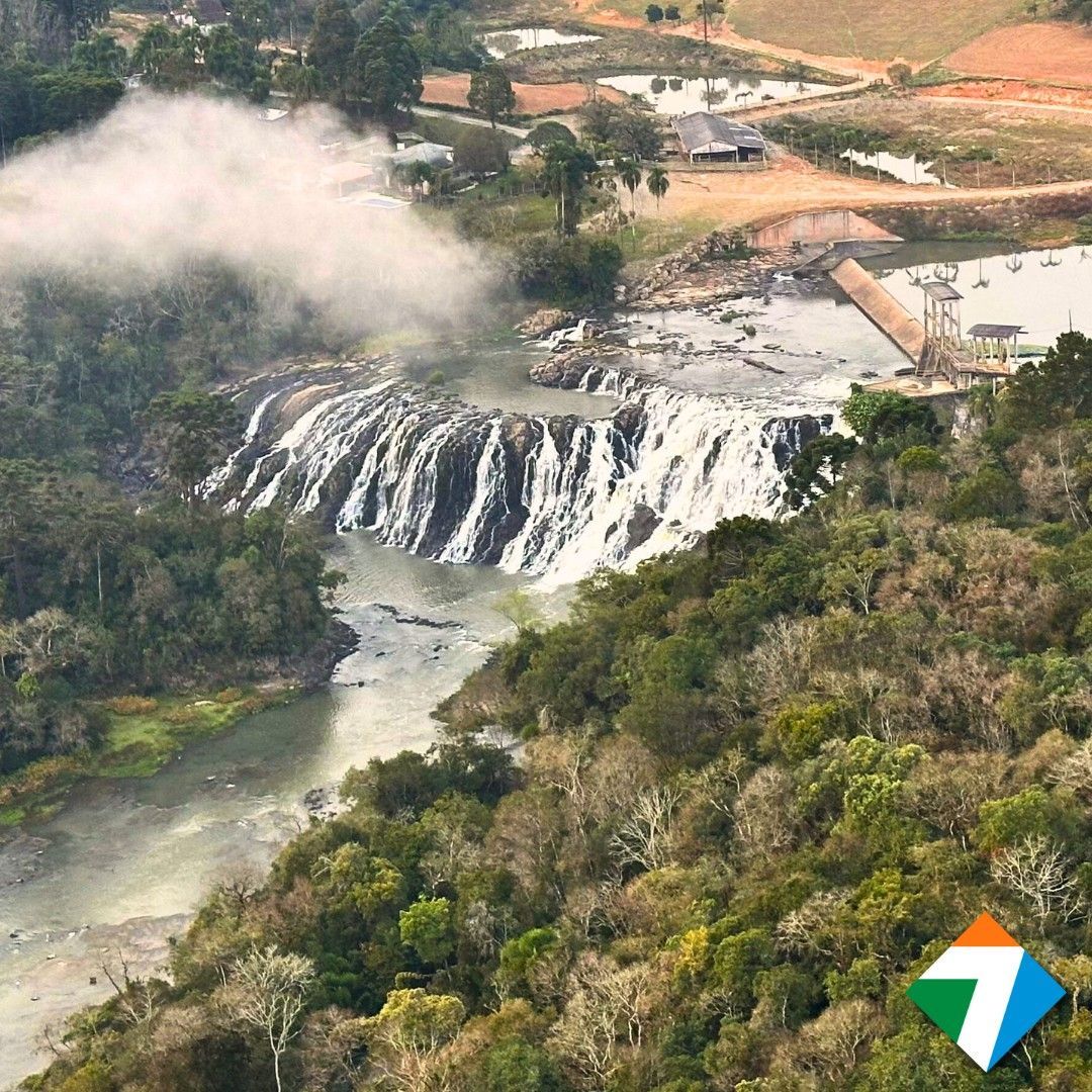 Cachoeira que deságua em um rio, cercada por árvores verdejantes.