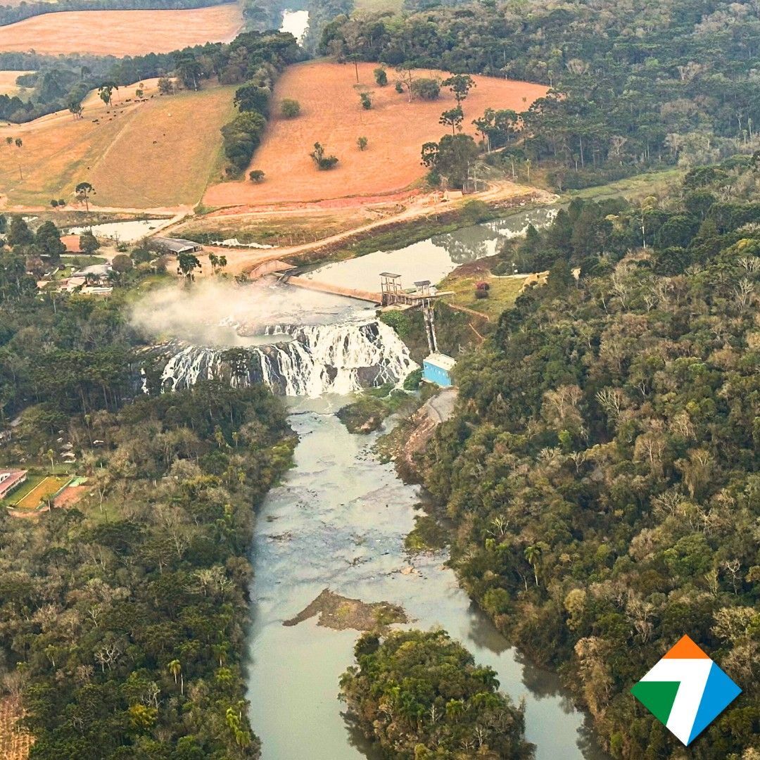 Vista aérea de um rio que deságua em uma cachoeira, cercado por árvores verdejantes e um campo na encosta.