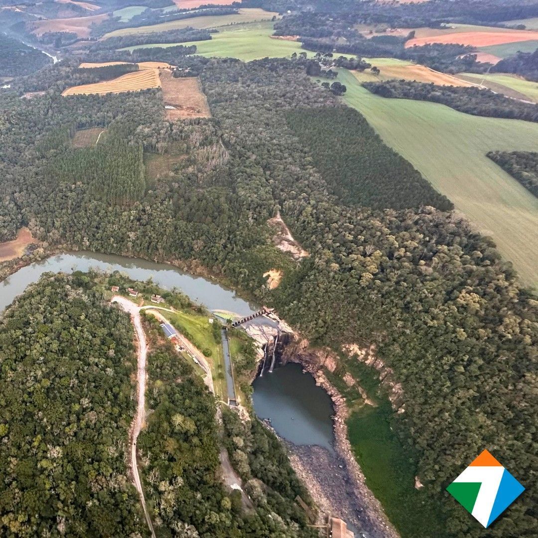Vista aérea de uma barragem e reservatório rodeados por floresta e terras agrícolas em um ambiente rural.