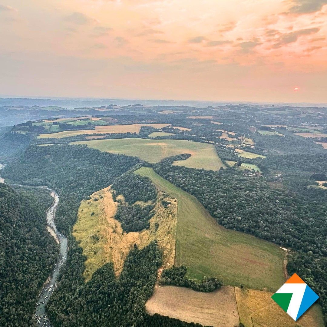 Vista aérea de uma paisagem com florestas, campos e um rio sob um céu enevoado ao pôr do sol.