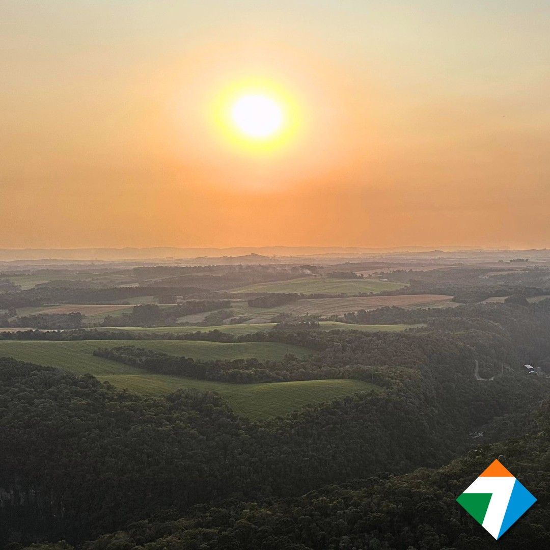 Pôr do sol sobre uma paisagem com florestas e campos, banhada por uma luz dourada.