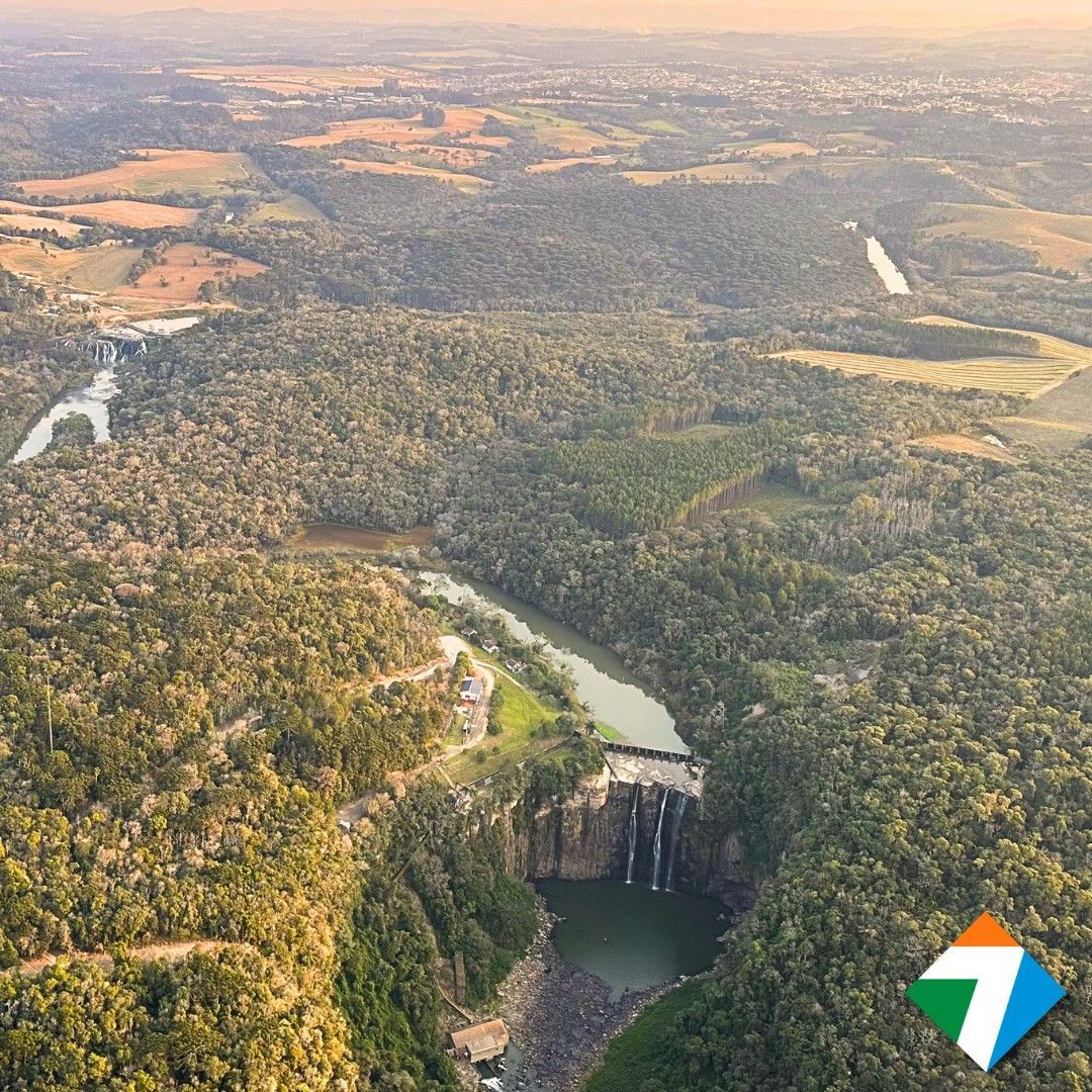 Vista aérea de uma exuberante floresta verde com um rio, uma represa e uma cachoeira na parte inferior.