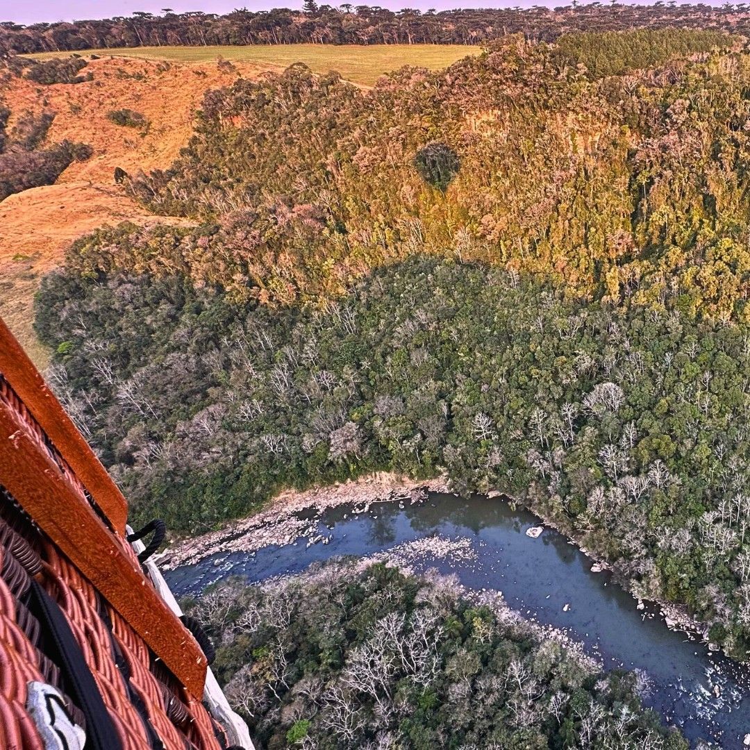 Vista aérea de um rio serpenteando por uma paisagem exuberante em tons de verde e bege, parcialmente encoberto por uma parte de um balão de ar quente.