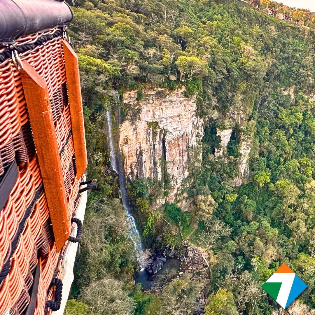 Vista da cesta de um balão de ar quente de uma cachoeira que despenca de um penhasco, cercada por uma exuberante floresta verde.